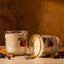 Two gold spiked cider candles with white wax and autumn-themed labels, one standing upright and one lying on its side, styled on a tabletop with cinnamon sticks and dried leaves against a warm textured background.